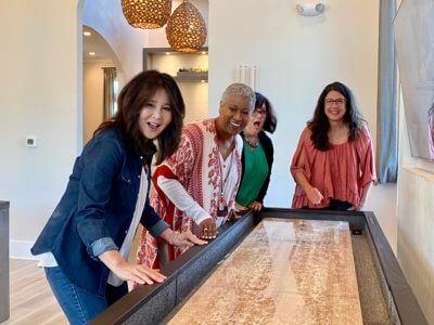 Four women standing at a shuffle board table watching one woman take a shot