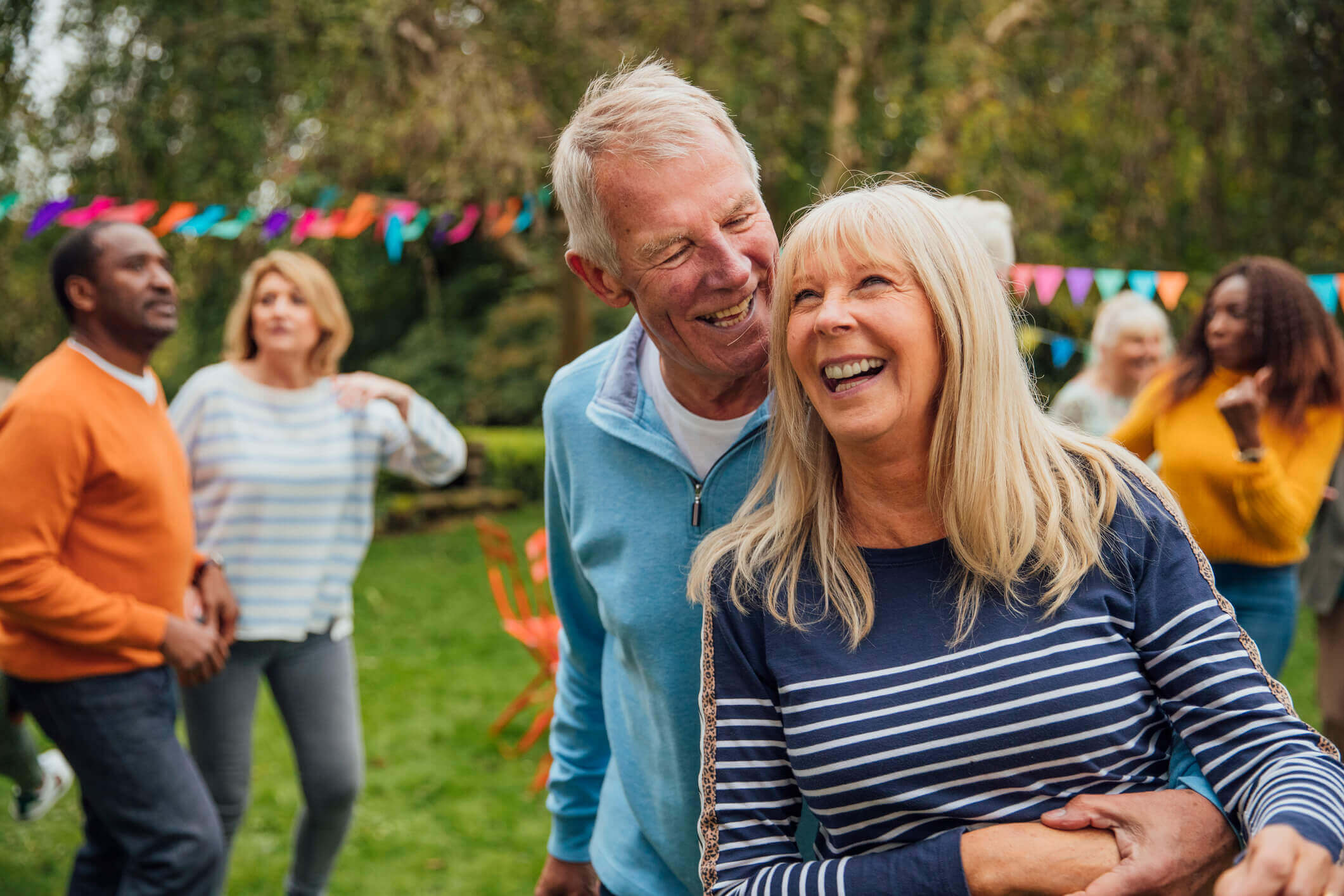 A man and his wife enjoy a cuddle together in the garden at an autumn tea party. Colorful bunting decorate the garden. Groups of people chat and dance in the background.
