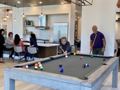 Two men playing pool with people sitting in the kitchen behind them talking