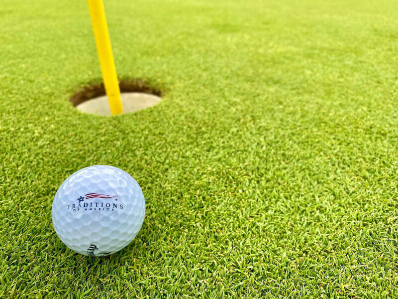 Golf ball with traditions of America logo next to the hole on a golf course